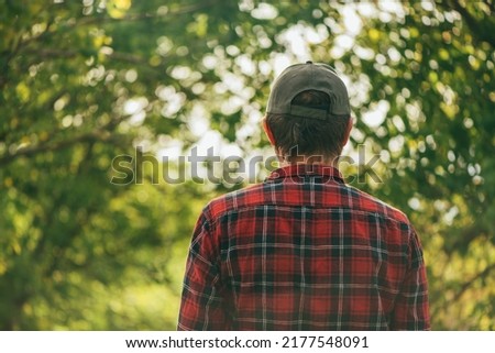 Rear view of male farmer wearing plaid shirt and trucker's hat standing in walnut orchard and looking at trees, selective focus Royalty-Free Stock Photo #2177548091
