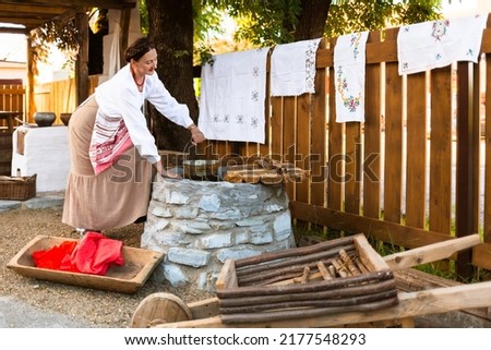 a woman in the national dress of an East European peasant Slavic Russian Ukrainian at a well with a rocker on a rural farmstead is collecting water in a bucket Royalty-Free Stock Photo #2177548293