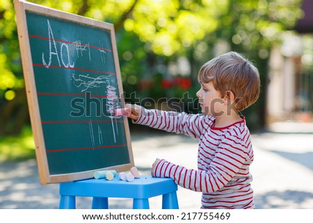 Cheerful kid boy at blackboard practicing writing letters, outdoor school or nursery