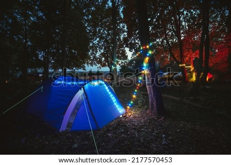 Night in the camp. Blue tent with colored lanterns in the forest. Travel and vacation in the wild nature. Chairs and barbecue. vehicle on the background.