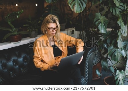 Millennial woman in classic eyewear checking received email message on modern netbook device using 4g internet at sofa, skilled female blogger chatting in social media during wireless networking