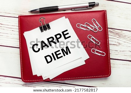 On a light wooden background, a burgundy pen and notebook. On the notebook has white paper clips and white paper with the text CARPE DIEM. Business concept