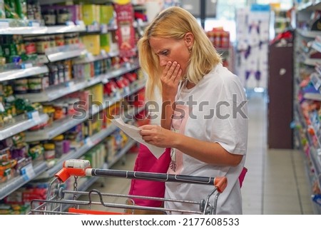 Woman looks shocked at a paper check in a grocery supermarket price increase and inflation. Upset woman in a supermarket viewing receipts looking at her shopping receipt and shocked about the price Royalty-Free Stock Photo #2177608533