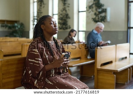 Young Christian black woman in casual dress kkeeping her eyes closed while sitting on bench in church and saying prayers to Lord Royalty-Free Stock Photo #2177616819