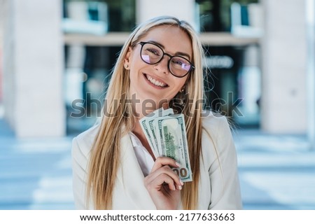 Excited blonde caucasian woman in glasses, white suite holding US dollar banknotes smiling wide, satisfied by profit against blurry buildings. Happy entrepreneur celebrates great deal, received cash. Royalty-Free Stock Photo #2177633663