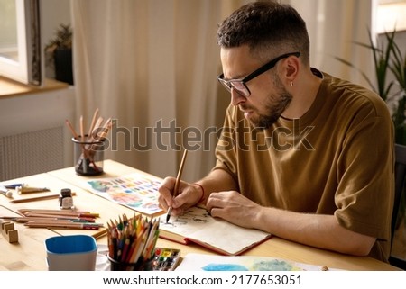 A young artist makes a brush sketch at his desk. Portrait of a creative person in his studio. Creative studio, lifestyle, the process of creating a work of art, the search for inspiration. Royalty-Free Stock Photo #2177653051