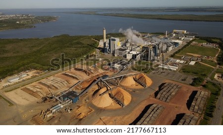 Aerial view of Paper Mill Factory with smoky chimneys front to River - Sawdust piles and pipes transporting wood trunks Royalty-Free Stock Photo #2177667113