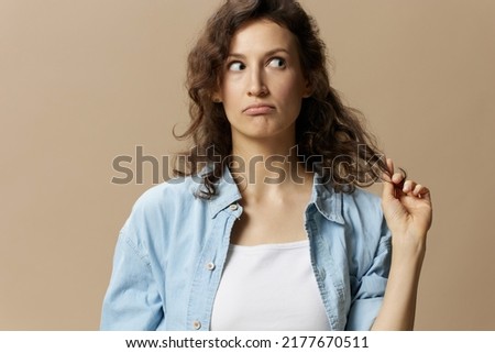 Confused pensive curly beautiful female in jeans casual shirt play with hair looks aside posing isolated on over beige pastel background. People Lifestyle emotions concept. Copy space
