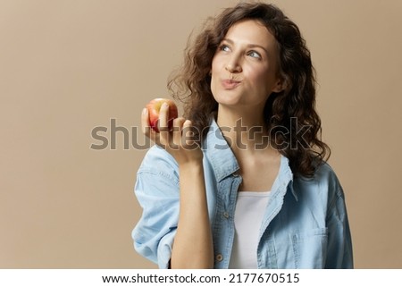 Funny happy cute curly beautiful female in jeans casual shirt chewing enjoy apple posing isolated on over beige pastel background. Healthy food. Natural eco-friendly products concept. Copy space Royalty-Free Stock Photo #2177670515