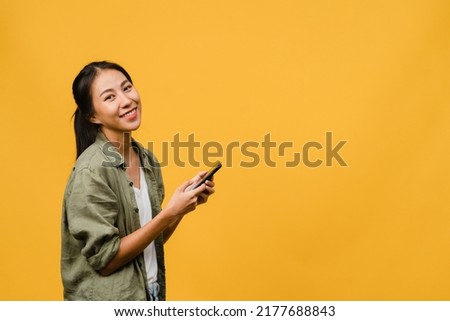 Surprised young Asia lady using mobile phone with positive expression, smile broadly, dressed in casual clothing and looking at camera on yellow background. Happy adorable glad woman rejoices success.