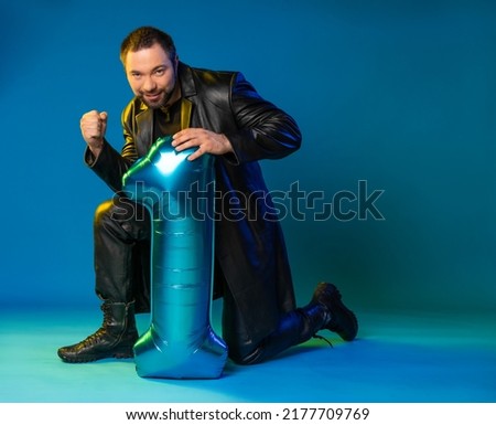 a man in a studio in a black leather coat knelt down and holds a large blue number one. wine background