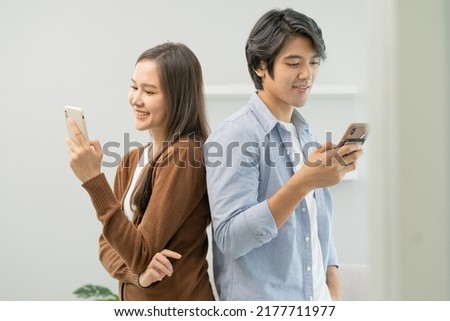 Infidelity, suspicion asian young couple love standing back to back, husband watching his wife using mobile phone, spying his girlfriend while woman typing a message, man distrust and jealousy at home Royalty-Free Stock Photo #2177711977