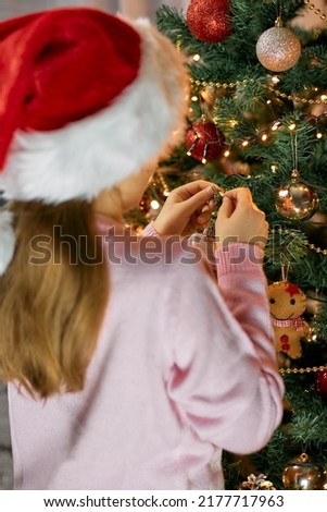 A girl in a Santa hat decorates a Christmas tree for the New Year or Christmas. Decorating. Waiting for the holidays. Traditions in preparation.