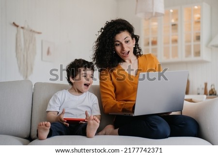 Amazed mother and son sitting on sofa at kitchen, watching cartoon by laptop, screaming together. Curly young woman spending time with little boy using gadgets. Happy family concept. Boy with phone. 