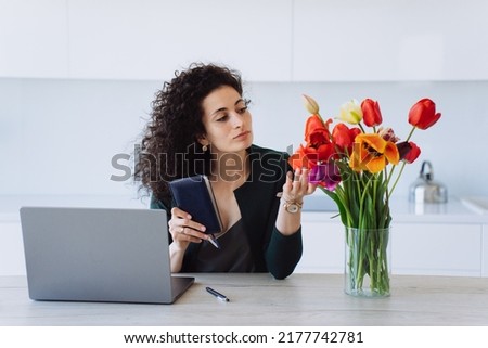 Pretty confident Italian woman in dark green suit looking at colourful tulips in vase, touching flowers sitting at table, needs a break. Curly businesswoman working at home. Remote working. Business.