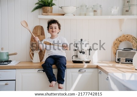 Playful little curly boy in white t-shirt and dark pants laughing sitting on a kitchen table, having fun, misbehaving at home. Spanish boy playing with wooden spoons at kitchen, kitchen stuff around.