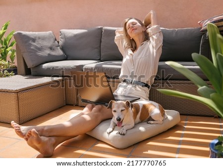 Blonde freelancer woman working remotely sitting on sunny terrace with laptop small senior cute dog Jack Russell terrier sleeps nearby. hands behind head. rest break from work. dreaming about vacation
