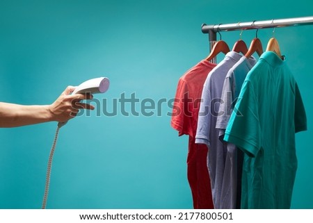 Males hand holds vertical steamer and using to iron colors t- shirts on blue background. Royalty-Free Stock Photo #2177800501