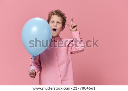 a beautiful cute little school-age boy stands with a blue balloon in his hand and emotionally poses with his mouth wide open. A photo on an empty pink background with space for an advertising insert