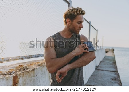 Young strong sporty athletic toned fit sportsman man in sports clothes warm up training using mobile cell phone app at sunrise sun dawn over sea beach outdoor on pier seaside in summer day morning.