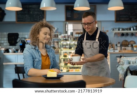 Caucasian male waiter with down syndrome serving a cup of coffee in the cafe to the client at the table Royalty-Free Stock Photo #2177813919