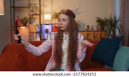 Young redhead child girl blogger taking selfie on smartphone selfie stick, communicating video call online with subscribers. Teen freckles kid at modern home apartment living room sitting on couch