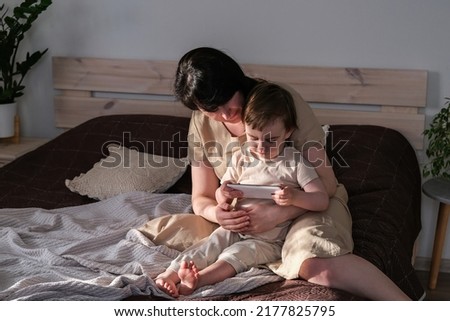 Mom and her little son sit on the bed and watching cartoons on a smartphone. The child watches a video on a digital gadget. Authentic lifestyle, real family home life