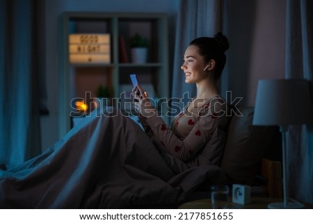 technology, bedtime and people concept - happy smiling teenage girl with smartphone and earphones sitting in bed at home at night