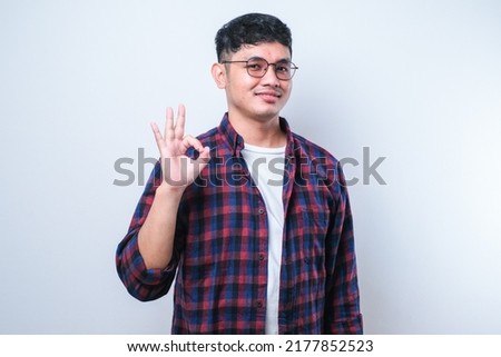 Young handsome Asian man wearing casual shirt and glassessmiling, doing ok sign, thumb up with fingers, excellent sign over white background