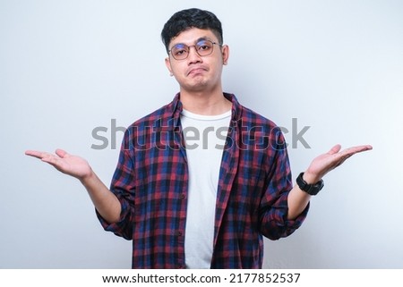 Young handsome Asian man wearing casual shirt and glasses over isolated white background clueless and confused expression with arms and hands raised. Doubt concept. Royalty-Free Stock Photo #2177852537
