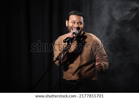 happy indian comedian sitting on chair and performing stand up comedy into microphone on black with smoke Royalty-Free Stock Photo #2177855701