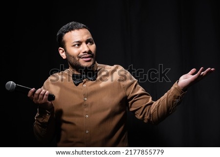 confused indian comedian in shirt and bow tie holding microphone and showing shrug gesture during monologue on black