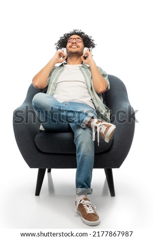 people and technology concept - happy smiling young man in headphones and glasses sitting in chair and listening to music over white background