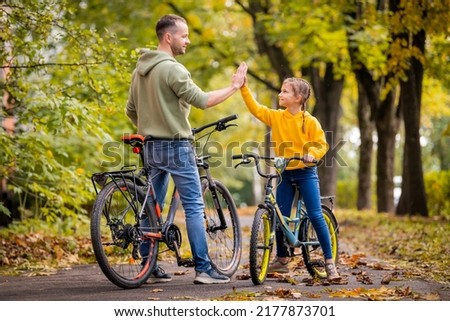 Happy father and daughter walk with bicycles in autumn park on sunny day.