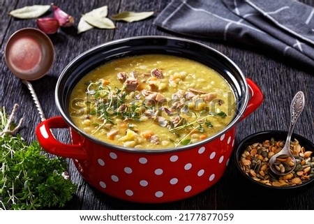 Finnish split pea soup Hernekeitto with ham on red pot with hot mustard and pumpkin seeds on dark wood table, horizontal view from above, close-up Royalty-Free Stock Photo #2177877059
