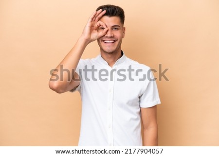 Young caucasian man isolated on beige background showing ok sign with fingers