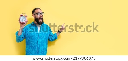 Portrait of smiling charismatic man holding pink alarm clock and pointing with finger at your text on yellow background. Funny promotion poster. Time to
