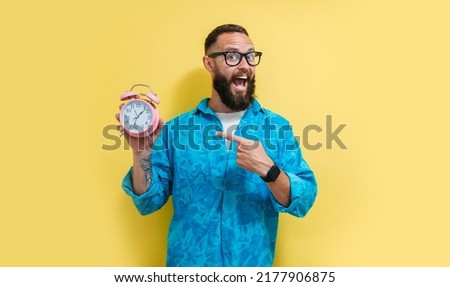 Portrait of smiling charismatic man holding pink alarm clock and pointing with finger at your text on yellow background. Funny promotion poster. Time to