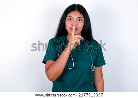 Doctor hispanic woman wearing surgeon uniform over white background  makes silence gesture, keeps finger over lips. Silence and secret concept.