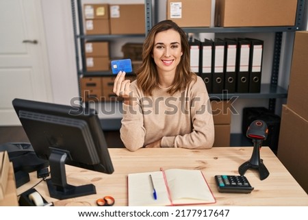Young woman working at small business ecommerce holding credit card looking positive and happy standing and smiling with a confident smile showing teeth 