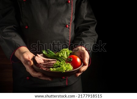 The chef serves a plate of sliced steak and vegetables. The concept of cooking on a dark background.