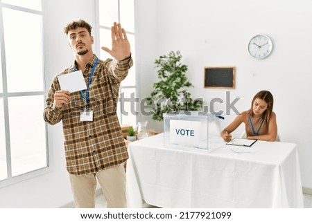 Young handsome man voting putting envelop in ballot box with open hand doing stop sign with serious and confident expression, defense gesture 