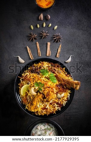 Chicken Dum Biryani table top photography on black-grey  background. Garnished with ingredients like garlic, star anise, Cinnamon, Green cardamom, and also with raita and red gravy in bowl. copy space Royalty-Free Stock Photo #2177929395