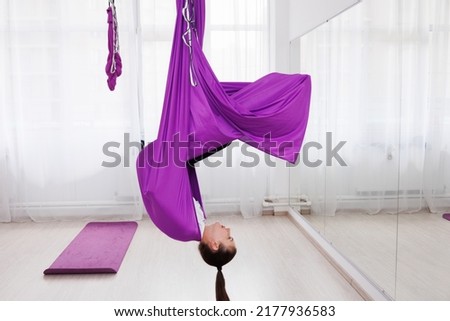 Young girl trainer of aero yoga holding  violet straps in a gym with large windows.  Pretty young woman standing in anti-gravity yoga hall. Mats laying on floor. Stretching and flexibility Royalty-Free Stock Photo #2177936583