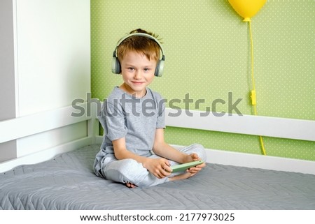 A preschool boy smiling sitting on the bed wearing wireless headphones and using mobile phone. Video chat, playing on cellphone, kids and technology concept.