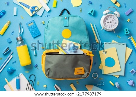 Back to school concept. Top view photo of open blue rucksack with plane shaped sharpener sticky note paper pens scattered school accessories drink bottle and alarm clock on isolated blue background
