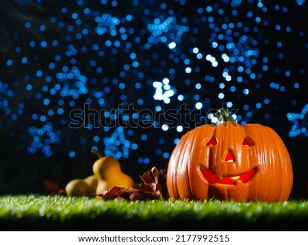 Halloween composition. Laughing orange pumpkin with carved face. glowing from within, autumn fruits, cones on green grass against a blue starry sky. Concept - Halloween.