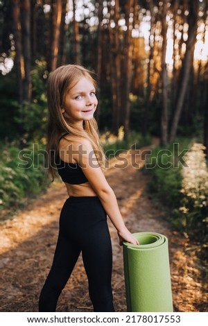 A stylish preschool girl, a professional athlete in a black summer suit stands with a green rug in her hands for gymnastics and yoga in the forest at sunset. Photography, concept.