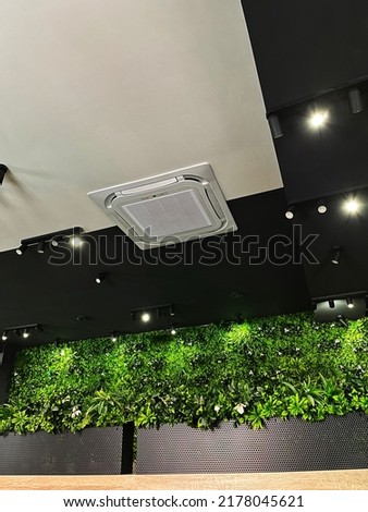 Ceiling air conditioner in a modern interior with ceiling lights, a green wall full of plants and wooden line in the bottom part of the picture.