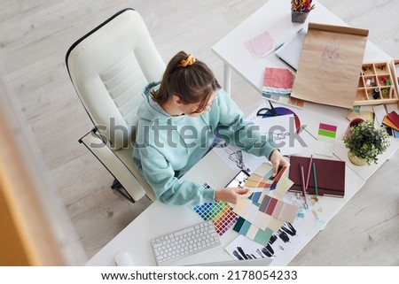 Above view of creative young designer sitting at table with massy sketches and choosing colors for new dresses